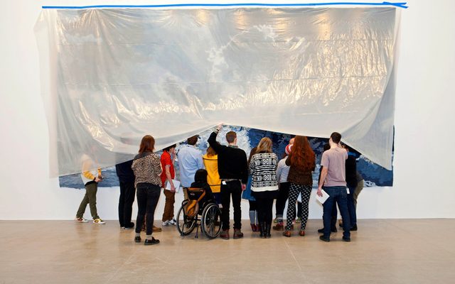 A group of people are lifting up a translucent cover to look at art hanging underneath it