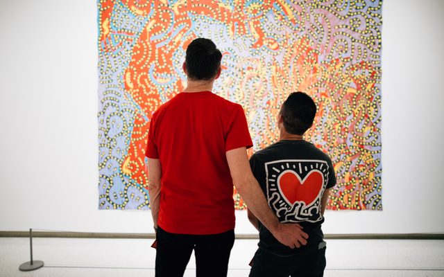 A taller adult has their hand on the lower back of another adult wearing a heart Keith Haring Tshirt, while looking at a displayed painting.