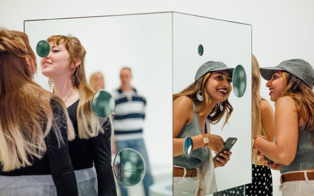 2 adults smiling and observing a mirrored-cube art installation