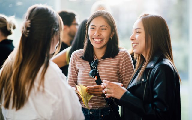 Three young women smile and chat together at an indoor social gathering or event.