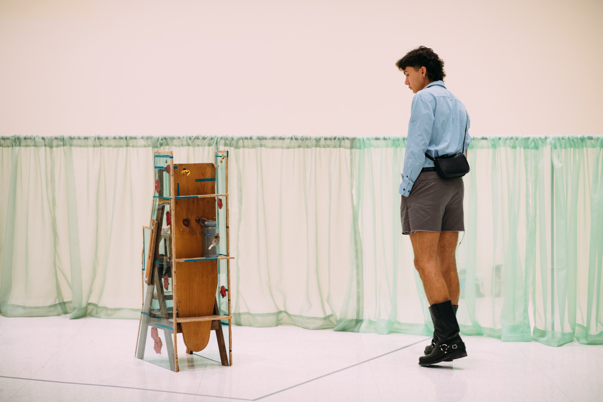 An adult walking around and looking at a stand-alone art piece, surrounded by sheer green low-hung curtain