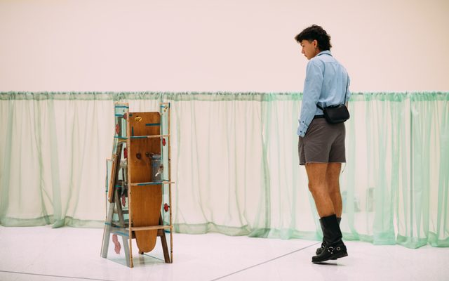 An adult walking around and looking at a stand-alone art piece, surrounded by sheer green low-hung curtain