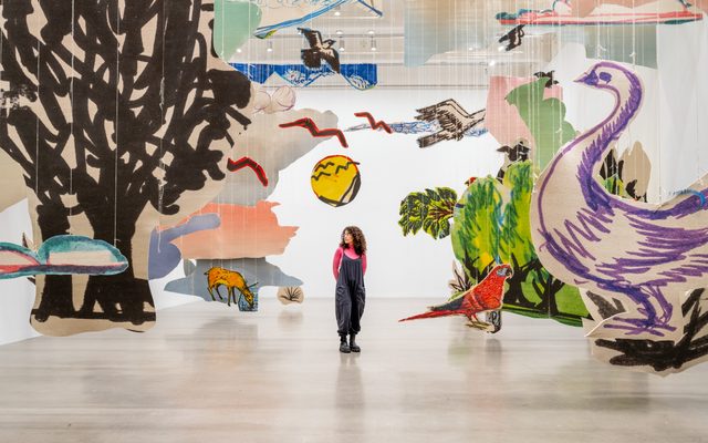An adult with a pink shirt and blue overalls stands in the middle of a large paper structure art installation