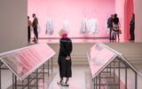 An adult with blonde and pink hair looks down at glass display in front of a clothing exhibit with a pink background