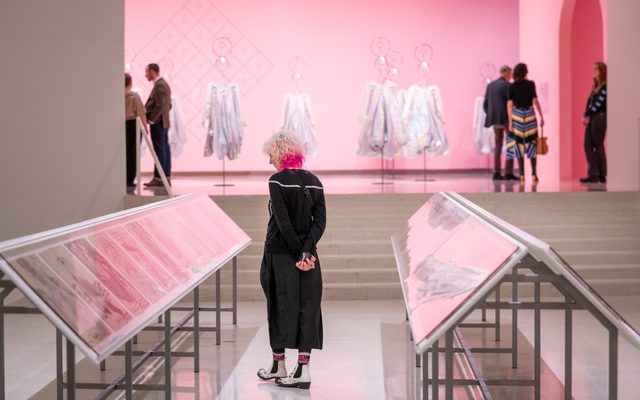 An adult with blonde and pink hair looks down at glass display in front of a clothing exhibit with a pink background