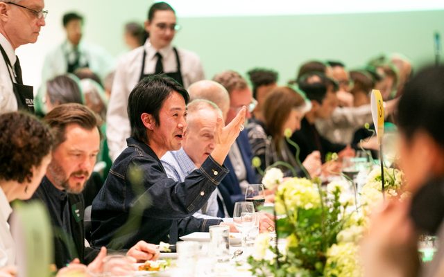 People seated at a long dinner table with flower center pieces, table signs, dinner and drinks with wait staff standing behind them