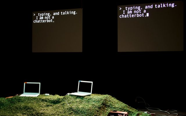 Two laptops are placed on a grassy surface, facing each other. Behind the laptops, two screens that display the text, 