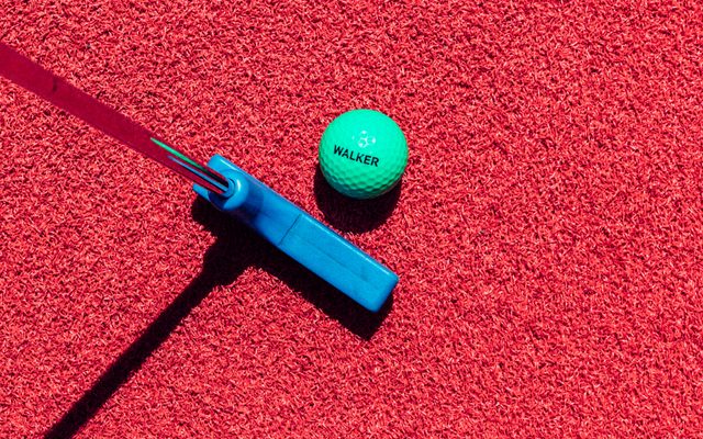 A green golf ball with the word WALKER printed in black, atop red turf grass next to a blue golf putter.
