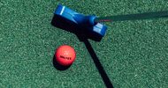 A red golf ball with the word WALKER printed in black, atop a turf grass next to a blue golf putter.