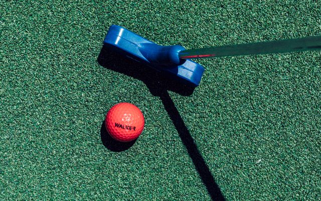 A red golf ball with the word WALKER printed in black, atop a turf grass next to a blue golf putter.
