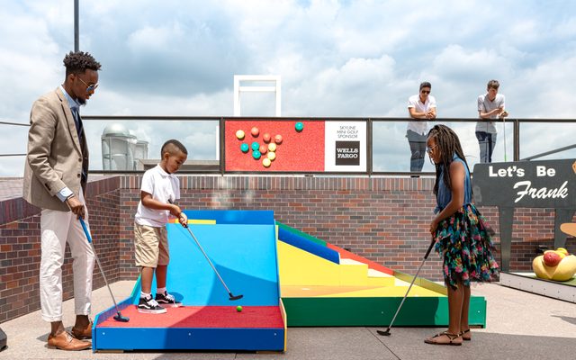 An adult and adolescent holding golf putters, watches an adolescent standing on a golf platform about to hit a gold ball with a putter.