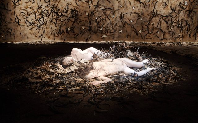 Live art installation of two adults painted in white and grime, laying on a pile of natural debris