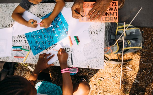 Two children and an adult make art from stencils on top of a bench in an outdoors sculpture garden.