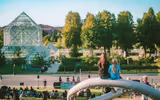 Two girls sit on a sculpture overlooking a park with people gathered and glass buildings in the background.