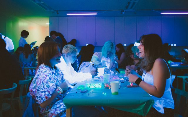 Peoples sitting at tables, making glow-in-the-dark crafts in a blacklit room