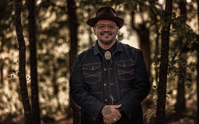 An adult with medium brown skin stands looking at the viewer in a forest. They are wearing a brown fedora and jean jacket.