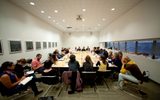 A group of adults sit and listen in a conference room at a table filled with papers and beverages