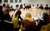 A group of adults sit and listen in a conference room at a table filled with papers and beverages