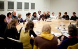 A group of adults sit and listen in a conference room at a table filled with papers and beverages