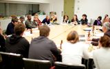 A group of adults sit and listen in a conference room at a table filled with papers and beverages