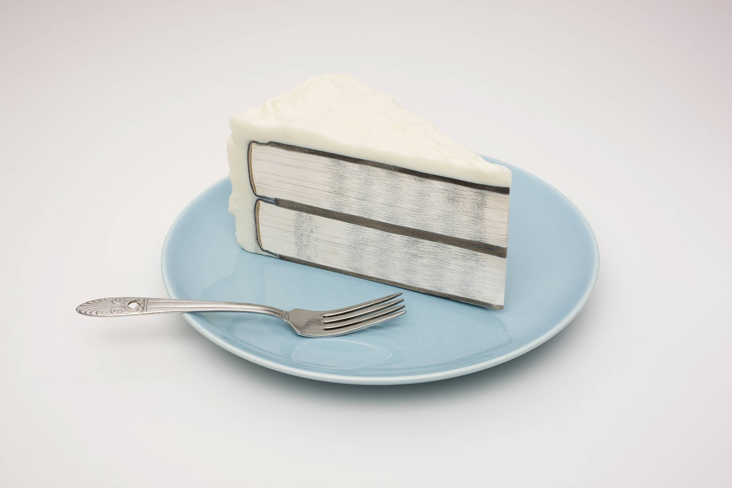 An art piece of a sliced white cake filled with two cut books that show the binding and pages, atop of a blue porcelain plate next to a fork