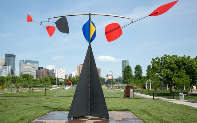 A large metal sculpture sits in a grassy lawn, a city view in the background. The sculpture has mobile-like arms with colorful objects on top.