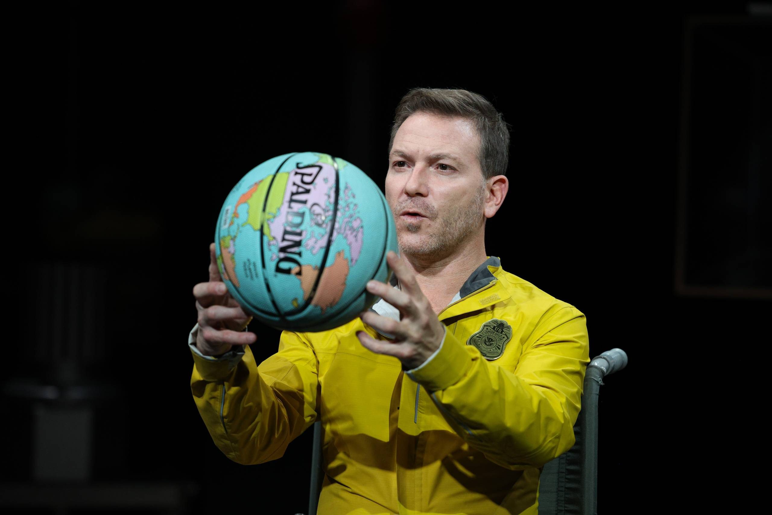 An actor in a bright yellow jacket holds up a basketball which features a print of the map of the world.