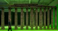 a horizontal image of a large room with columns made from packed earth sitting on a chromakey green surface that sits inches off of the floor. The blurred silhouette of a person walks past in the foreground.