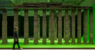 a horizontal image of a large room with columns made from packed earth sitting on a chromakey green surface that sits inches off of the floor. The blurred silhouette of a person walks past in the foreground.