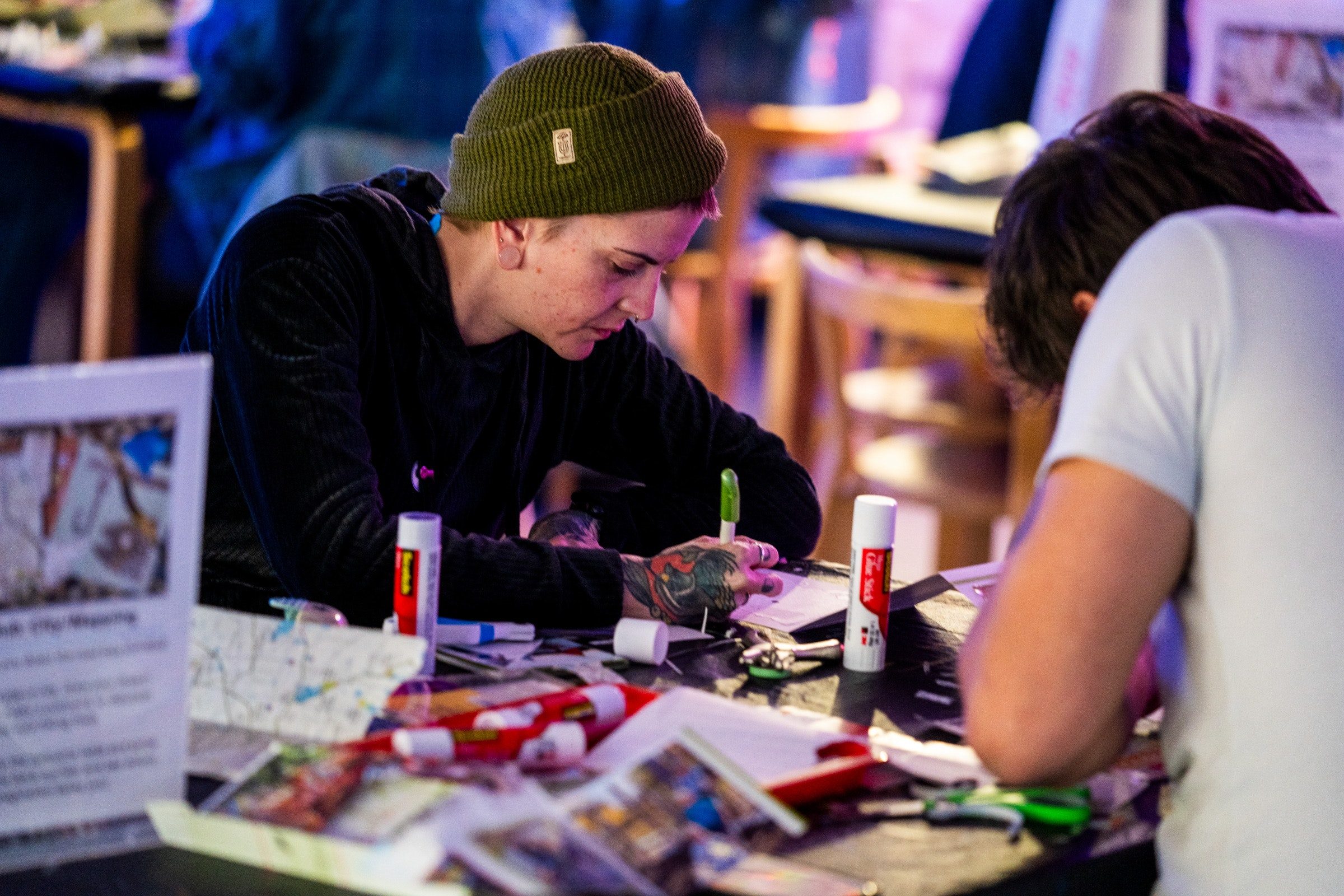 Two adults work on artwork with markers while seated at a table.
