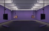 A purple-walled gallery with two suspended screens showing a woman, each facing opposite directions. Purple benches are placed on the floor beneath each screen. Ceiling lights illuminate the space.
