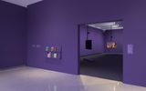 A minimalist art gallery with purple walls, a small bookshelf displaying colorful books, and two video screens in a darkened room, one showing a person gesturing. The floor is glossy and white.