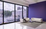 A person sits on a white sofa in a modern room with a purple wall, large windows featuring abstract patterned film, and minimalist white furniture on a gray rug.