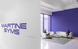 A gallery entrance with Martine Syms on the wall. Two people sit on white sofas in a lounge area with a purple accent wall and large window, reading or working quietly.