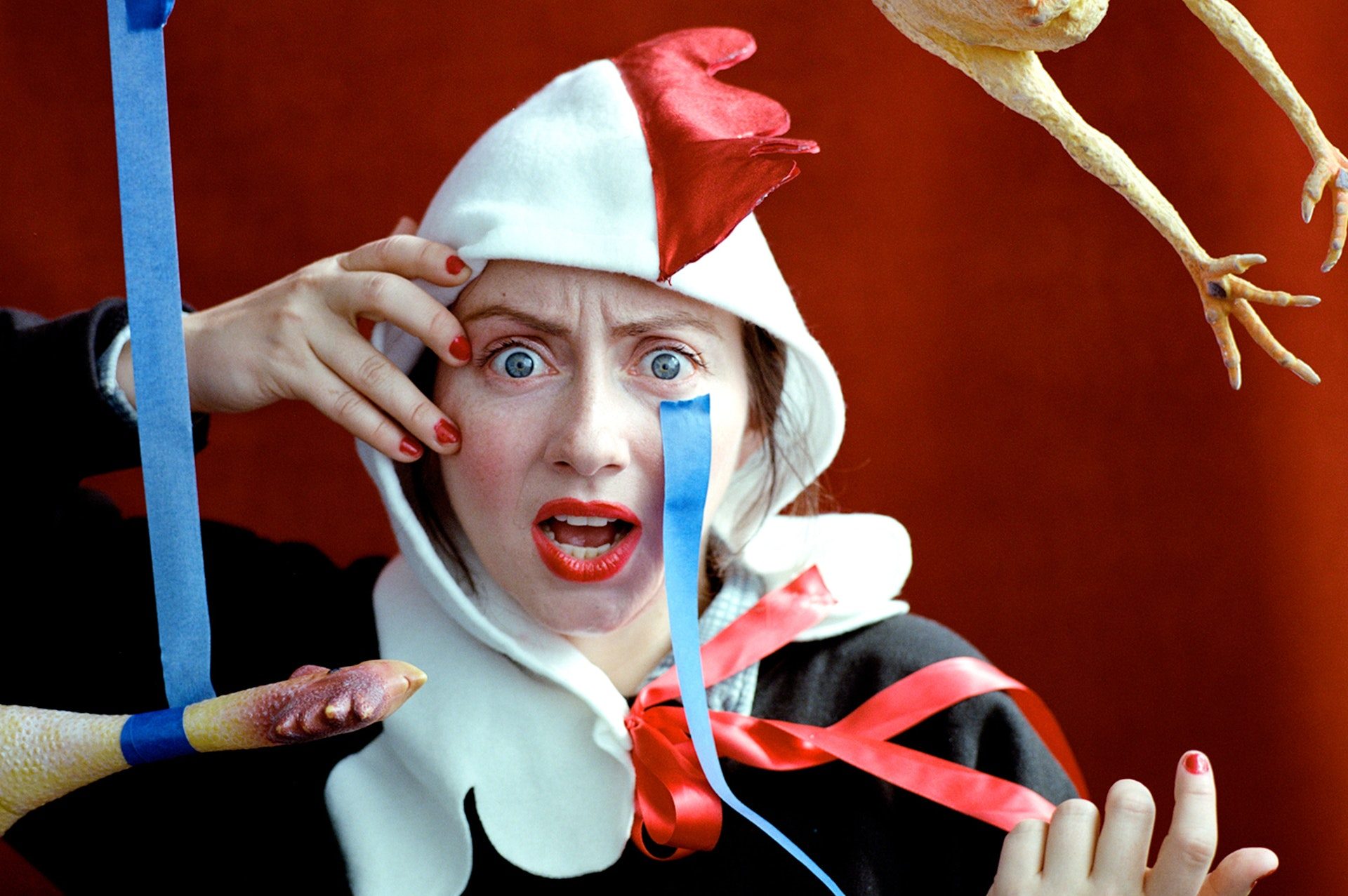 A light-skinned woman wearing a rooster head hood stares in shock at the viewer.