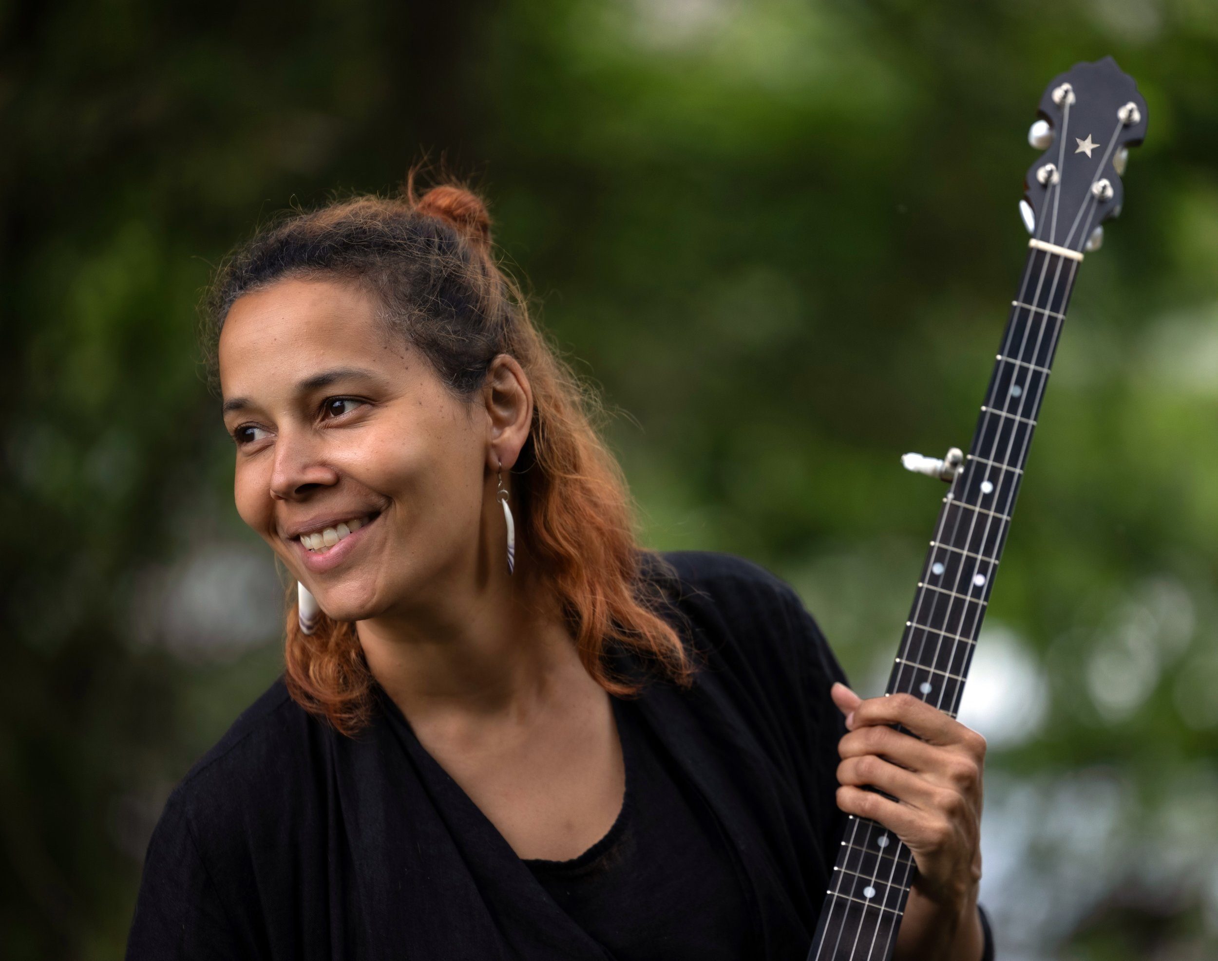 A woman with warm brown skin and copper-tinted hair smiles while holding a black banjo decorated with a star on its headstock.