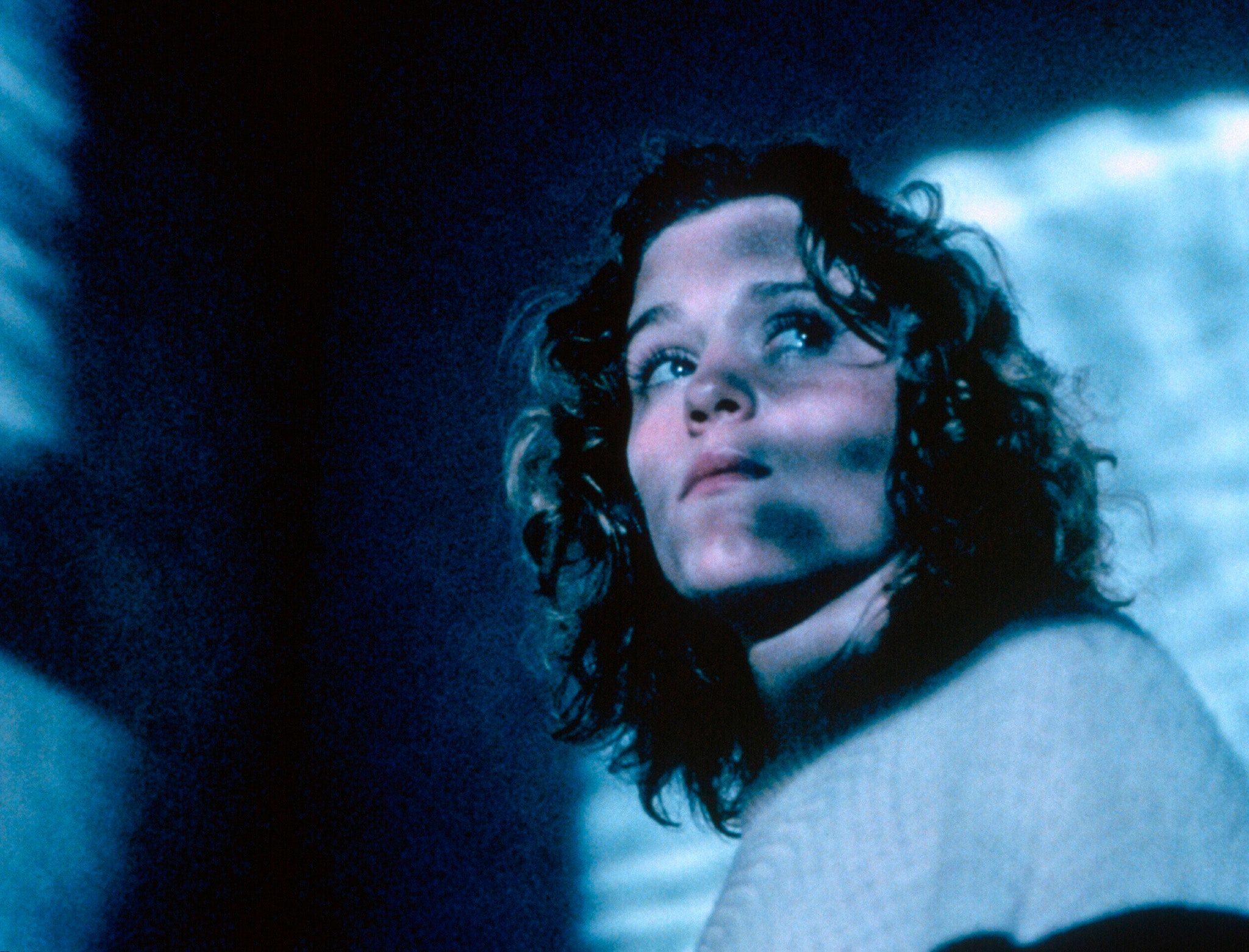A woman with short, curly dark hair looks upward, her face lit by cool blue light and broken by soft shadows, as if seen at night through a window or windshield. The background is dark and indistinct, creating a tense, cinematic mood.