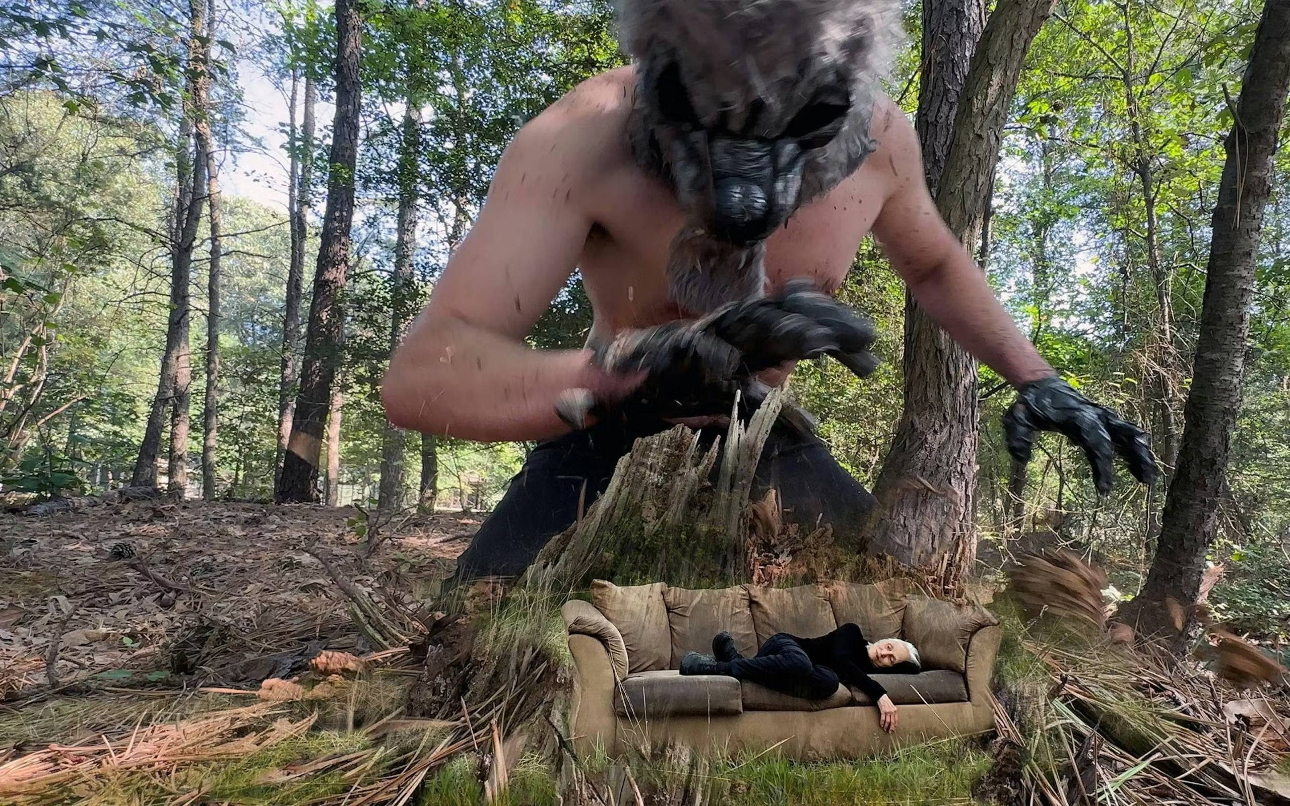 A woman in black clothing lays on a brown couch in the woods, composited with a man in the gloves and mask of a bear creature, towering over her.