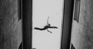 A blurred, black-and-white image shows a person mid-air between two tall buildings, appearing to leap or fall, with arms and legs extended against a bright sky background.