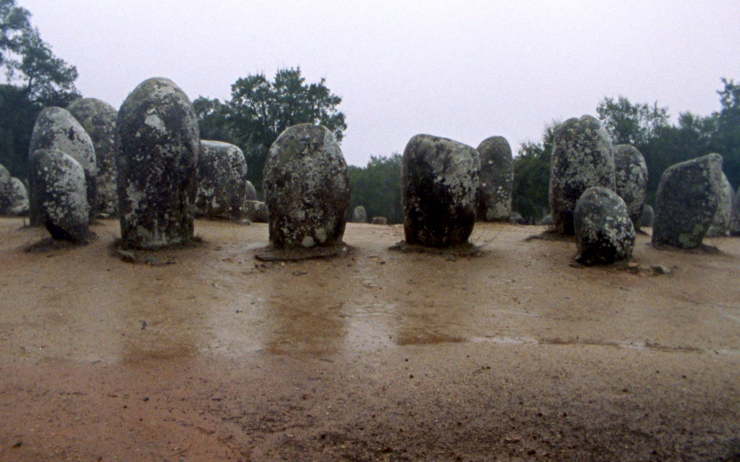 A monolith of 12 massive rocks sitting a strange circle on sand. Even more large rocks, partially obscured, can be seen in the background.