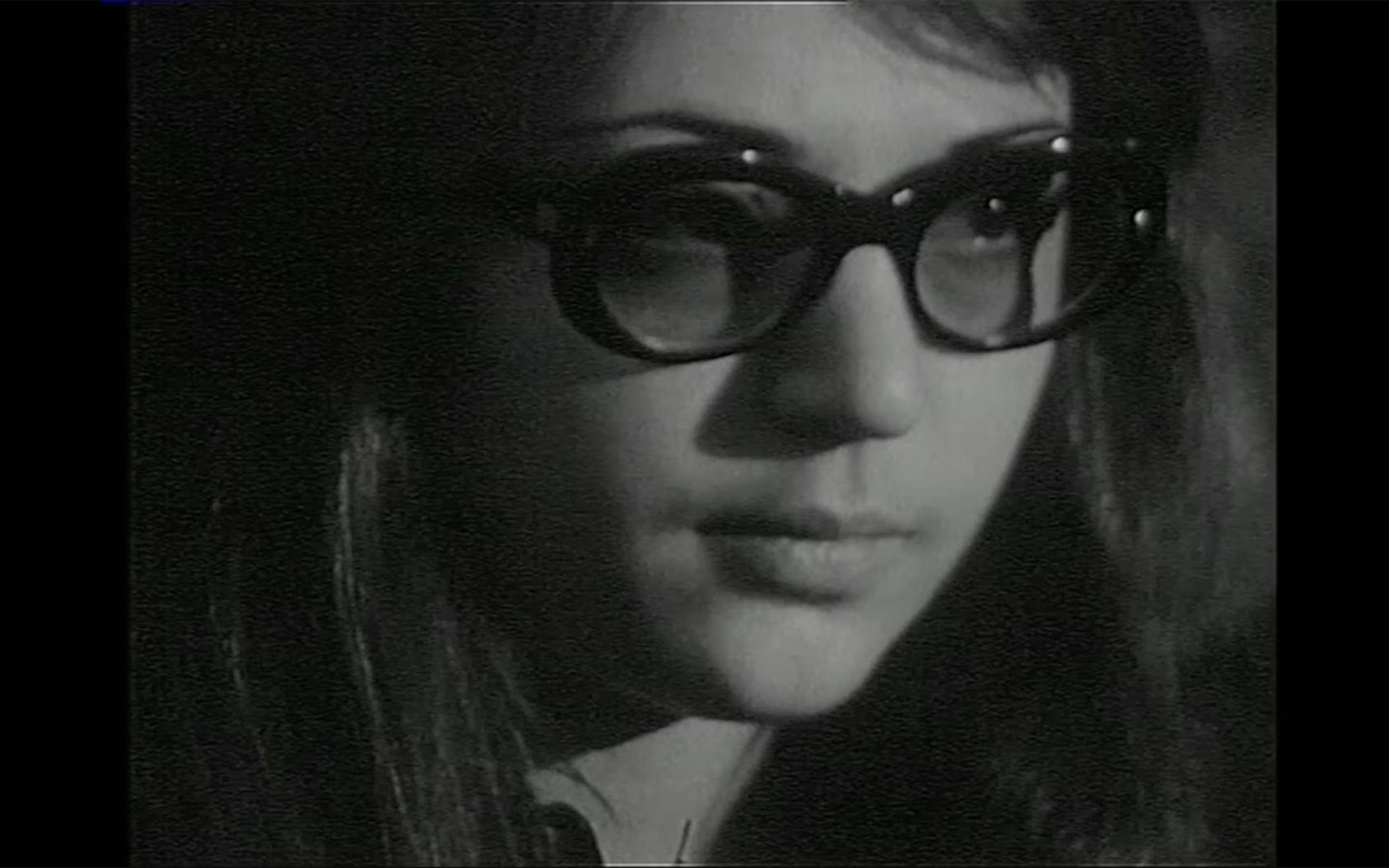 Black and white still of a close cropped shot of a girl's face with round plastic glasses and long straight hair, looking off to the right.
