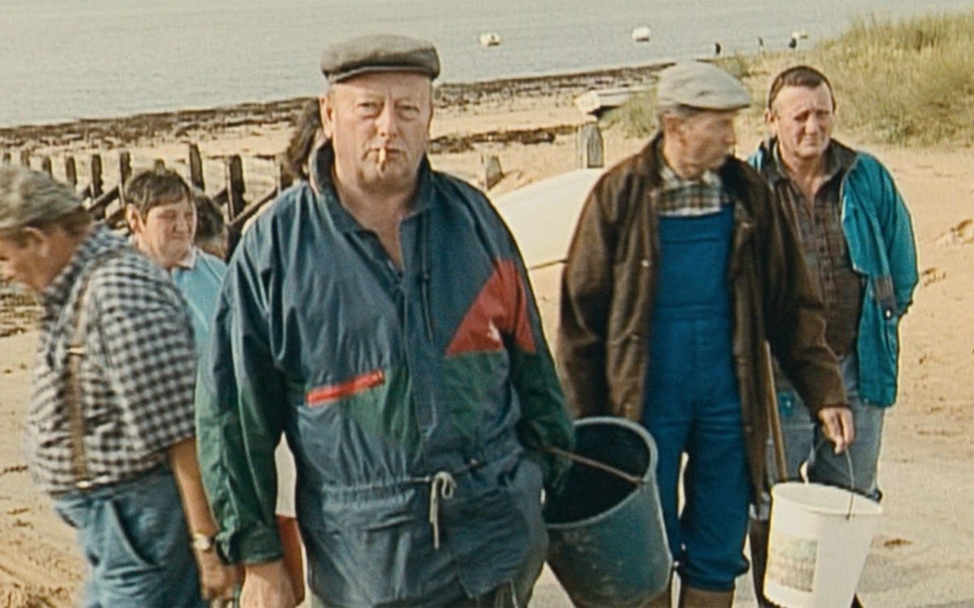 A handful of men in work clothes holding buckets walk together at the beach. One looks directly at the camera.