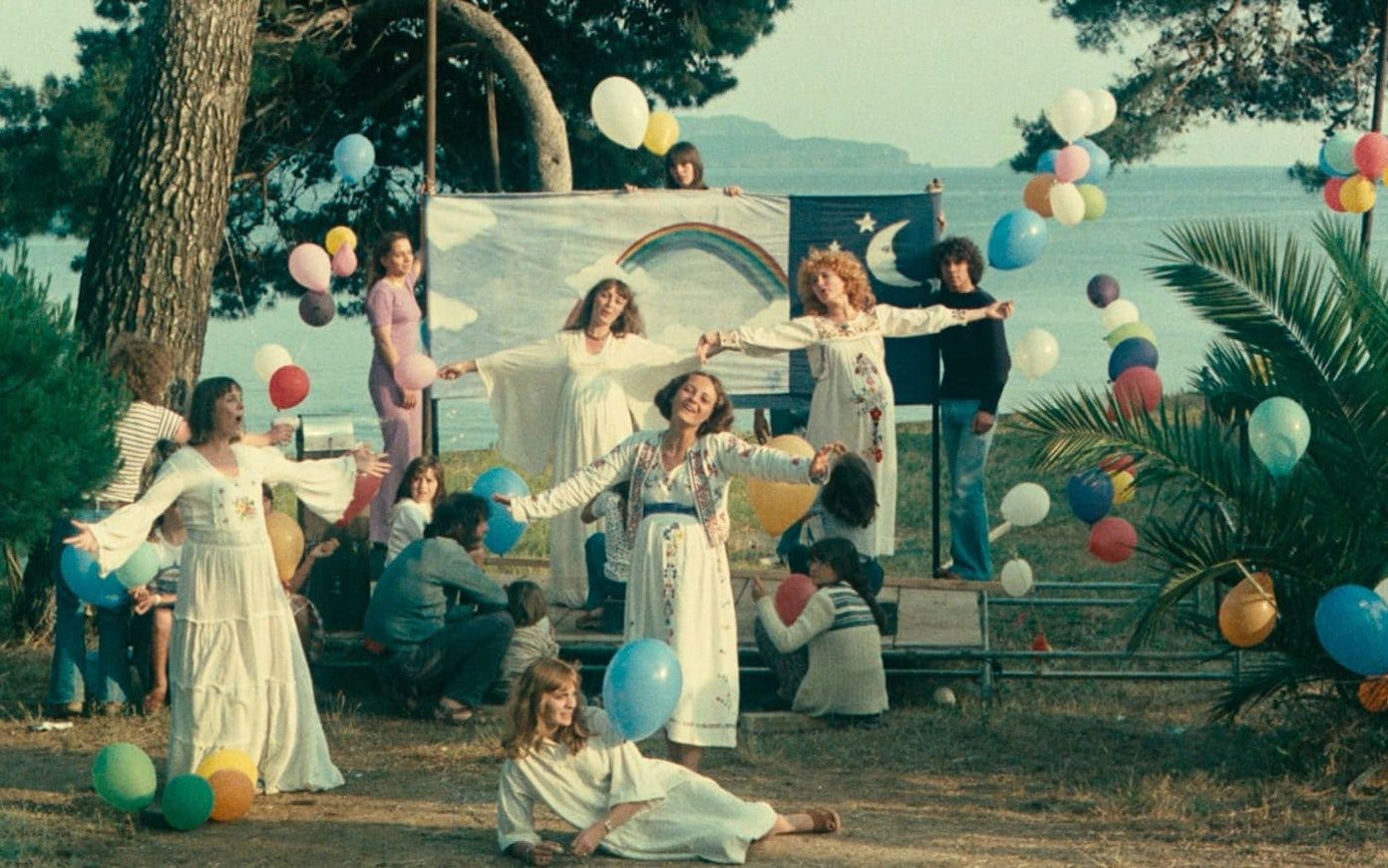 Musical scene with pregnant women in long dresses singing with arms out, surrounded by colorful balloons and a backdrop, with the sea in the background.