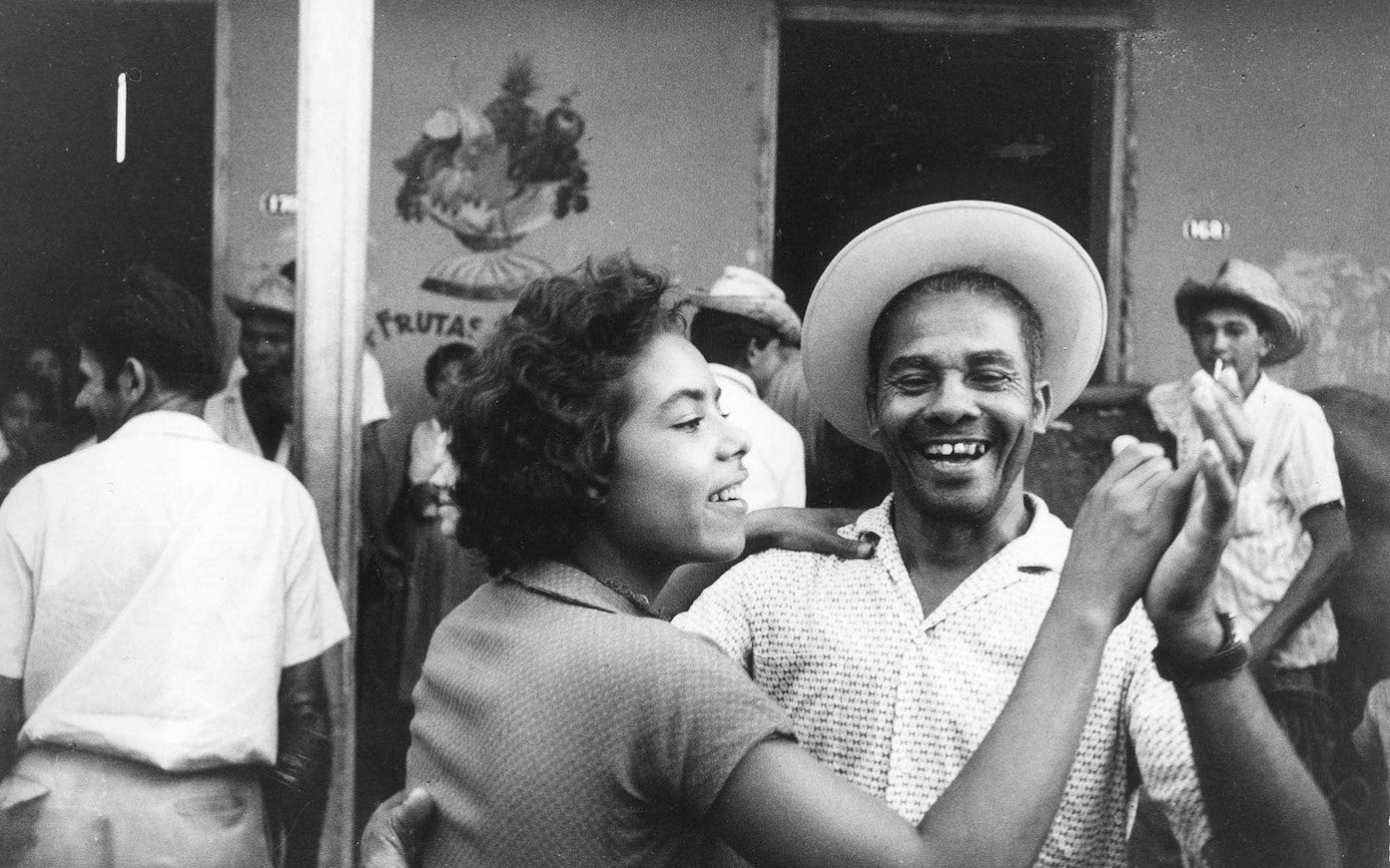 Black and white still of a man and woman with medium-dark skin dancing together outdoors and smiling, among a crowd of others.