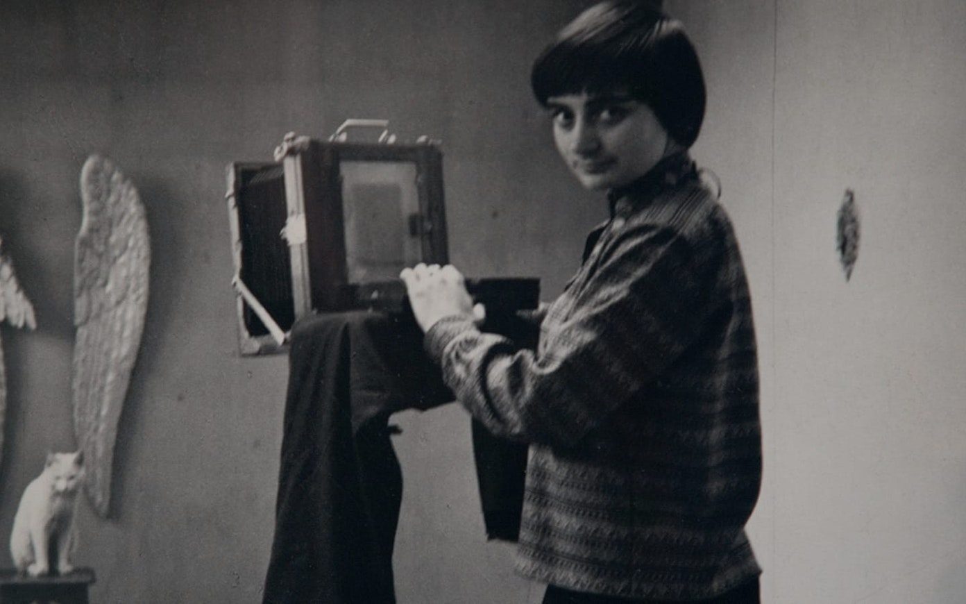 Black and white still of a young Agnes Varda looking at us from behind a large format camera aimed toward a white cat and wings hanging on the wall behind it.