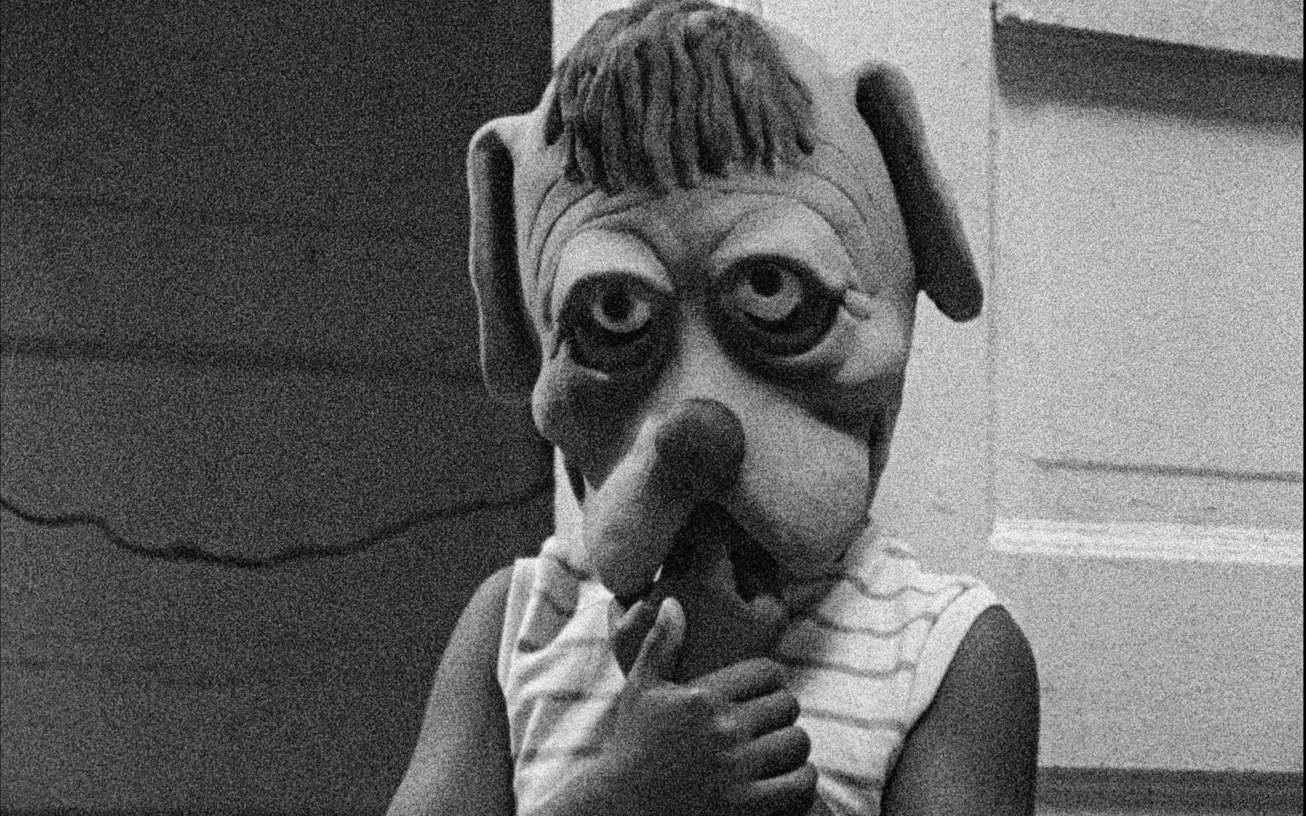 Black and white still of a kid in a striped shirt wearing an oversized mask of a cartoony dog head. The kid holds their hands up to their mouth.