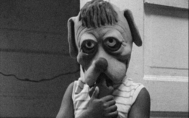 Black and white still of a kid in a striped shirt wearing an oversized mask of a cartoony dog head. The kid holds their hands up to their mouth.