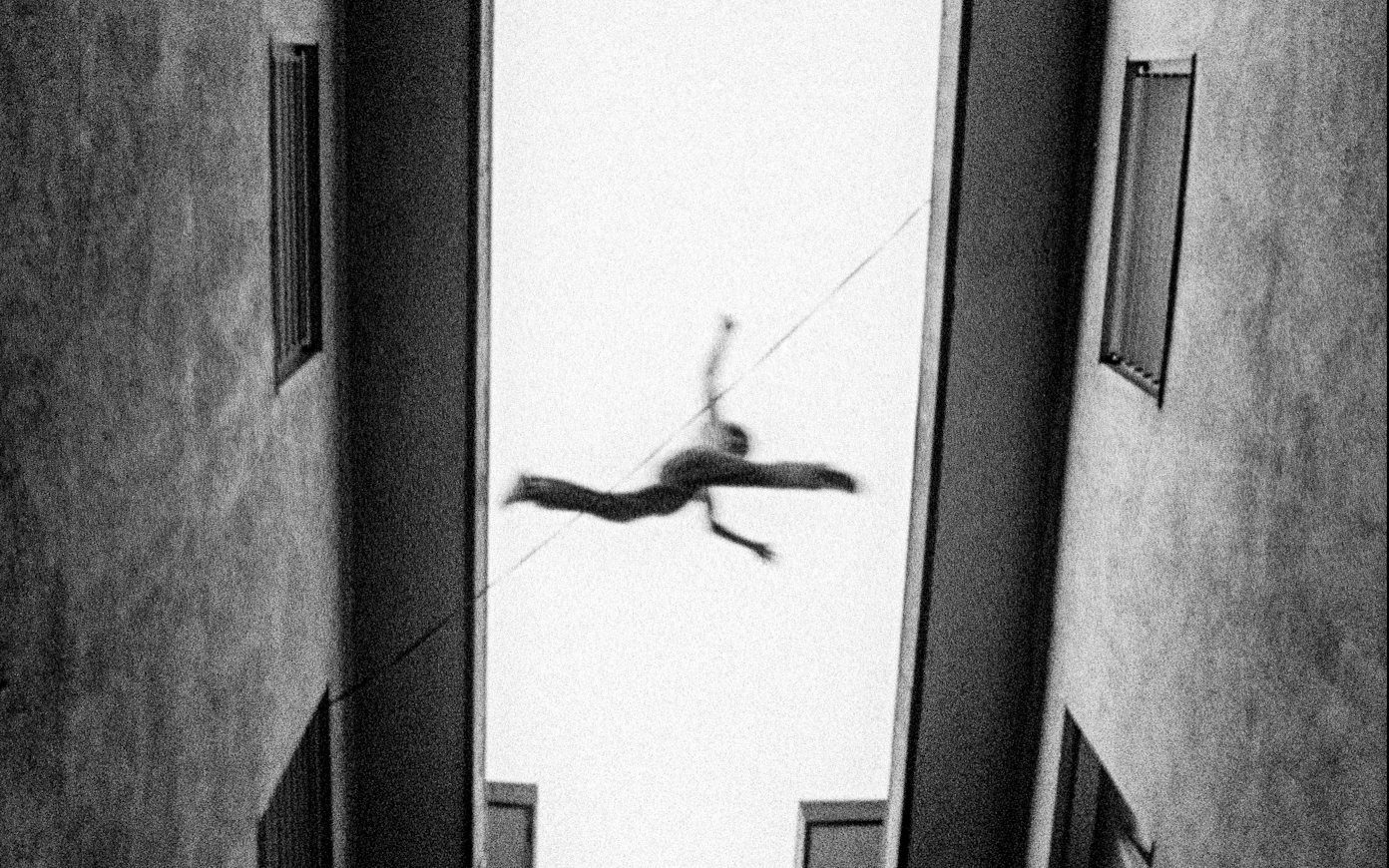 Black and white still shot from below of a young person leaping between the roofs of two buildings. They are mid-jump, not touching either side.