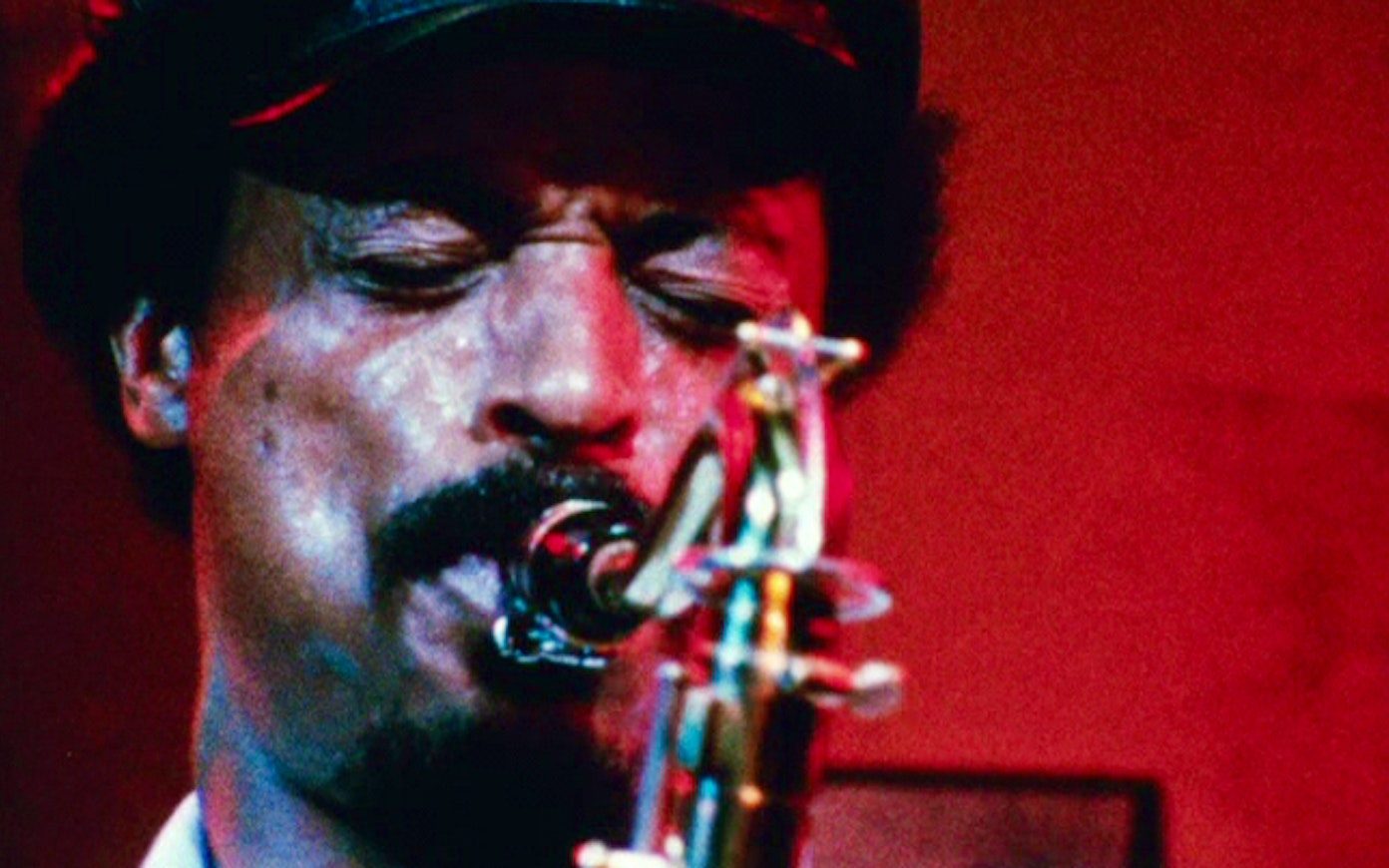 Very close still of a man playing a saxophone with his eyes squeezed shut, surrounded by red light.