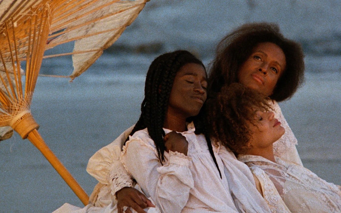 Three women in white dresses sit leaning against each other with content looks, on a beach with a sun umbrella leaning to the left side.